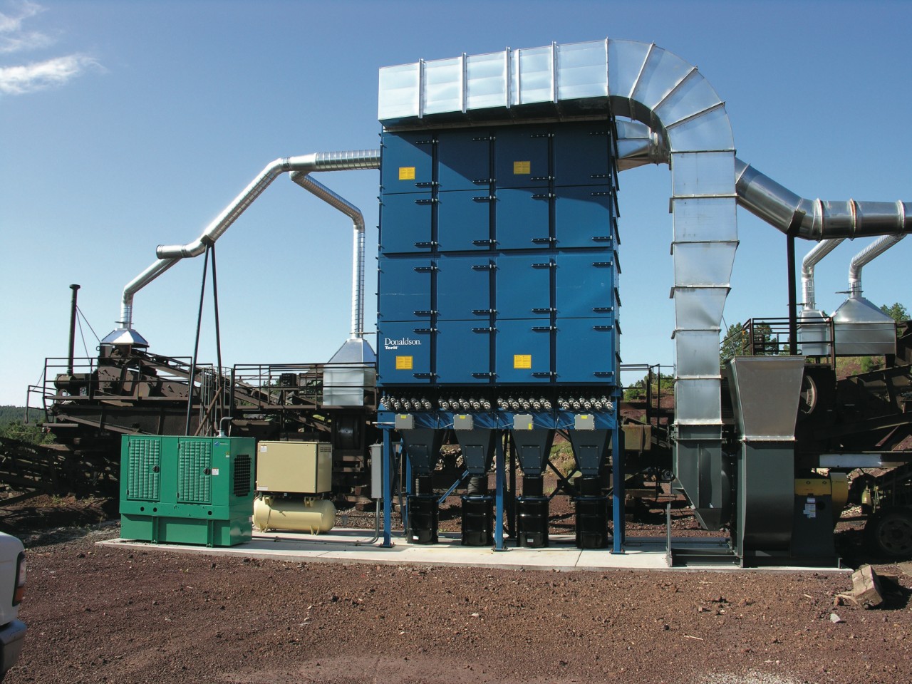 Un colector de polvo con filtro de mangas Torit® Dalamatic® de Donaldson centralizado, con conductos hacia los puntos de generación de polvo en los puntos de transferencia del transportador. Dado que este tipo de filtro de mangas limpia sus filtros con aire comprimido, la instalación necesitaba instalar un compresor de aire (aparato verde) y un secador de aire comprimido (equipo marrón) junto al colector.